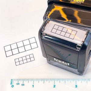May include: A black Trodat Printy 4913 stamp with a grid pattern is shown next to a ruler. The stamp is on a white surface with two printed grid patterns. The ruler shows inch measurements.