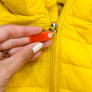 May include: A yellow puffer jacket with a zipper closure. A hand is holding a red security tag attached to the zipper.