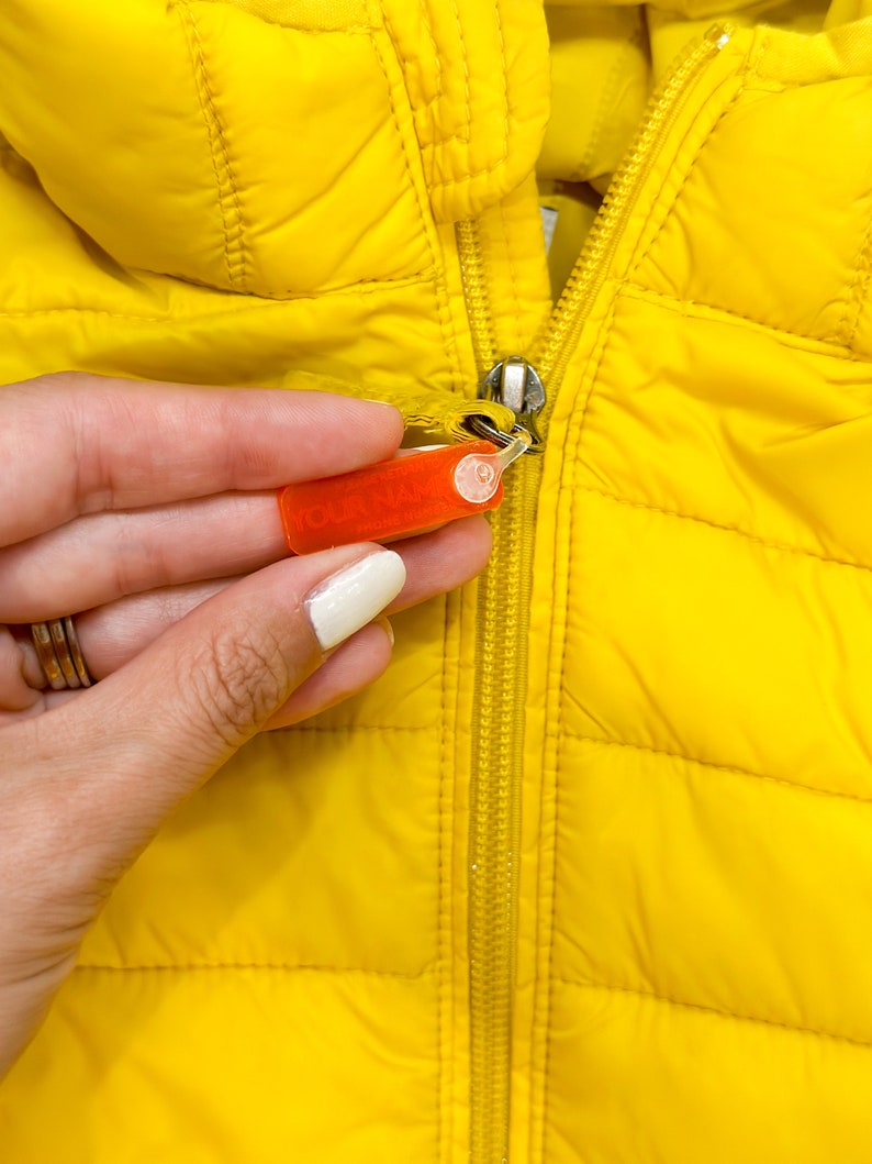 May include: A yellow puffer jacket with a zipper closure. A hand is holding a red security tag attached to the zipper.