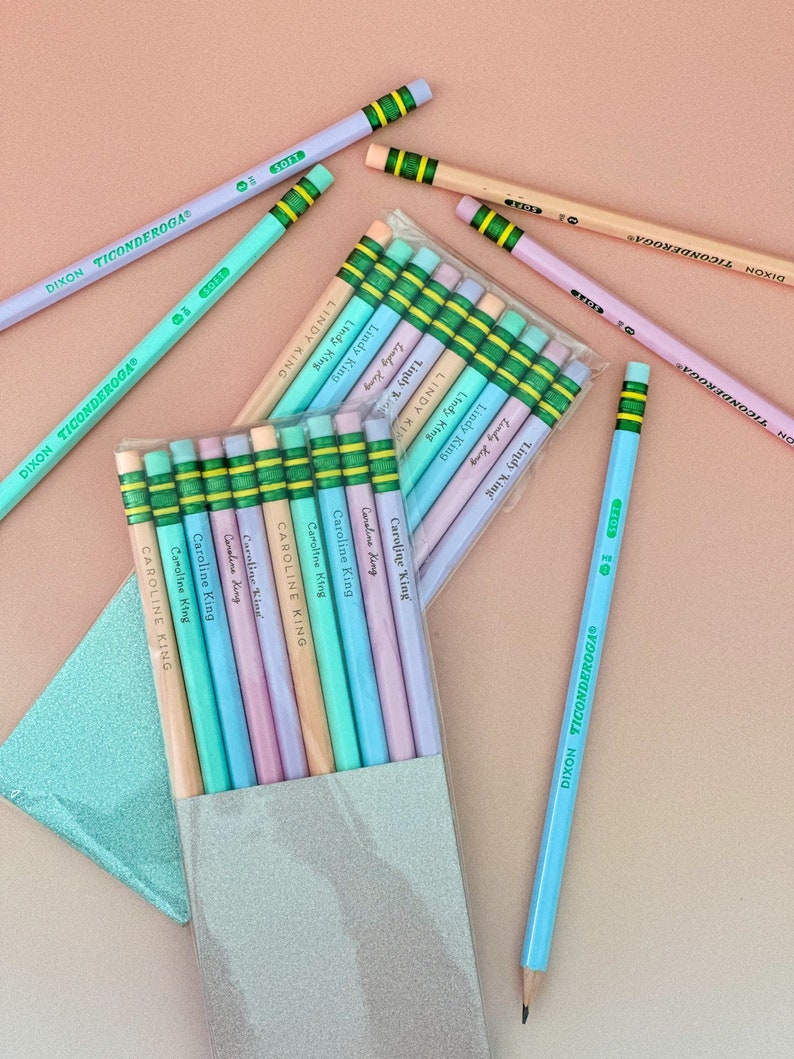 May include: Pastel-colored pencils, including light blue, lavender, and peach, are scattered on a pink surface. The pencils are labeled "DIXON TICONDEROGA" and "SOFT." Some pencils are packaged and labeled "Lindy King" and "Caroline King."