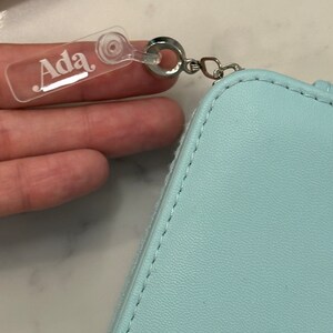 May include: A light blue zippered pouch with a clear plastic tag that says "Ada" attached to a silver ring.