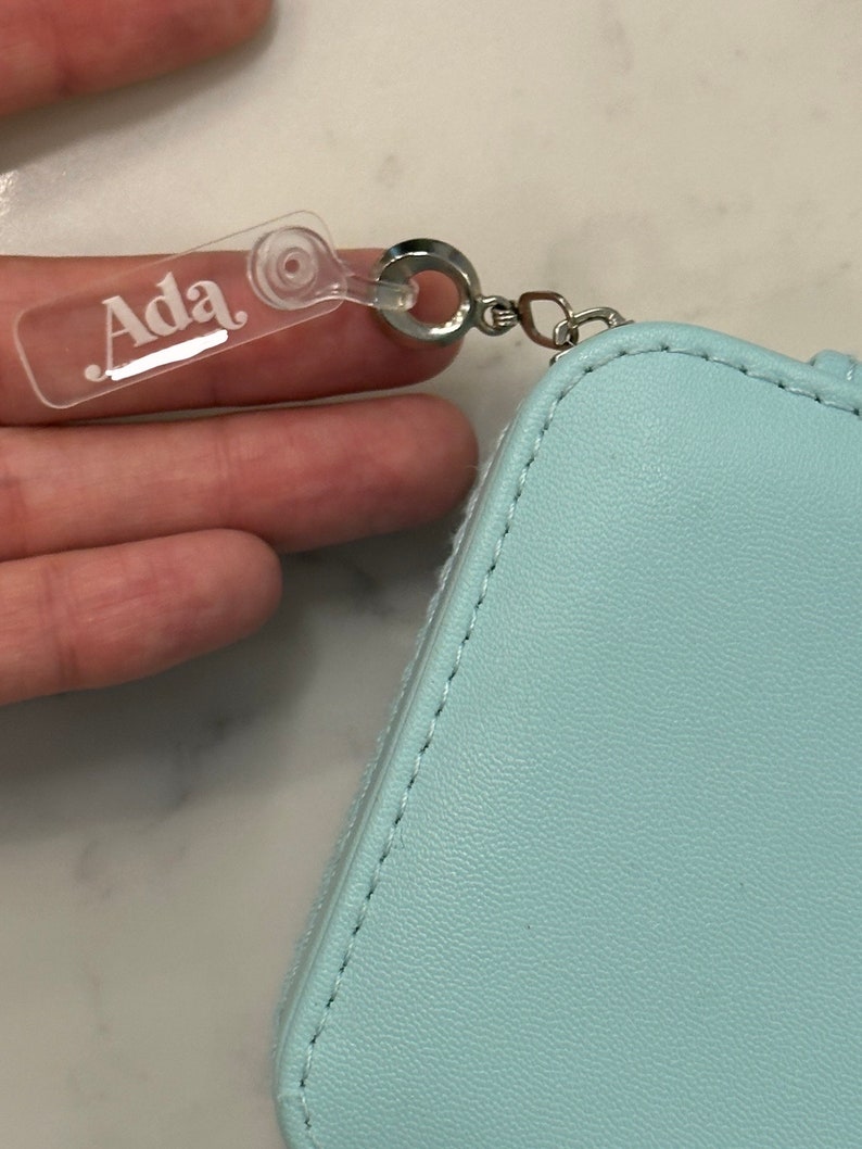 May include: A light blue zippered pouch with a clear plastic tag that says "Ada" attached to a silver ring.