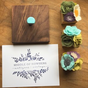 May include: A wooden block stamp with a floral design. The stamp is labelled "MIDDLE OF NOWHERE antiques + vintage". The image also shows three felt flower arrangements in small wooden boxes.