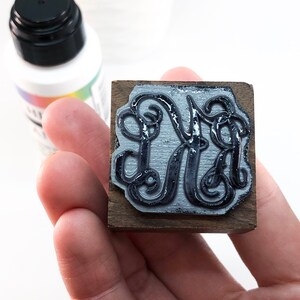 May include: A wooden stamp with a black ink pad featuring a decorative monogram of the letter "M" in a script font.
