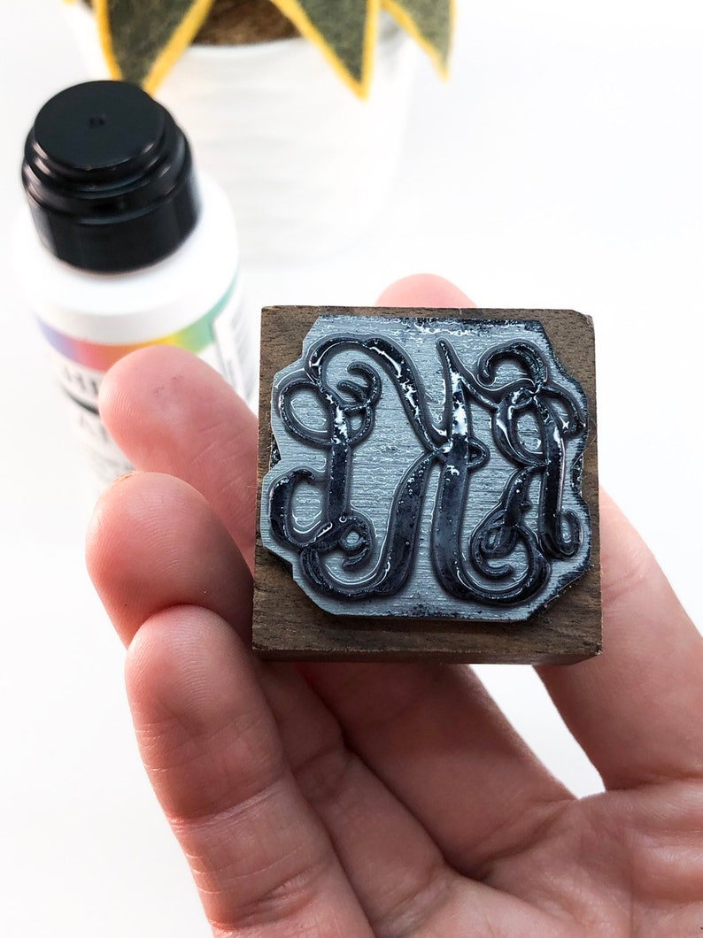 May include: A wooden stamp with a black ink pad featuring a decorative monogram of the letter "M" in a script font.