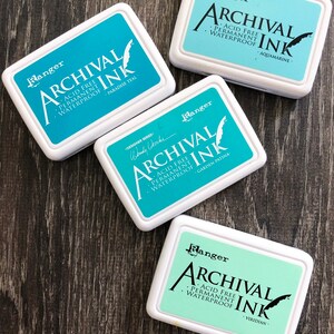 Peut inclure: Quatre tampons d'encre avec des couvercles blancs et de l'encre bleue et verte. Les tampons d'encre sont &eacute;tiquet&eacute;s "Ranger Archival Ink" et chacun a une couleur d'encre diff&eacute;rente : Paradise Teal, Aquamarine, Garden Patina et Viridian.