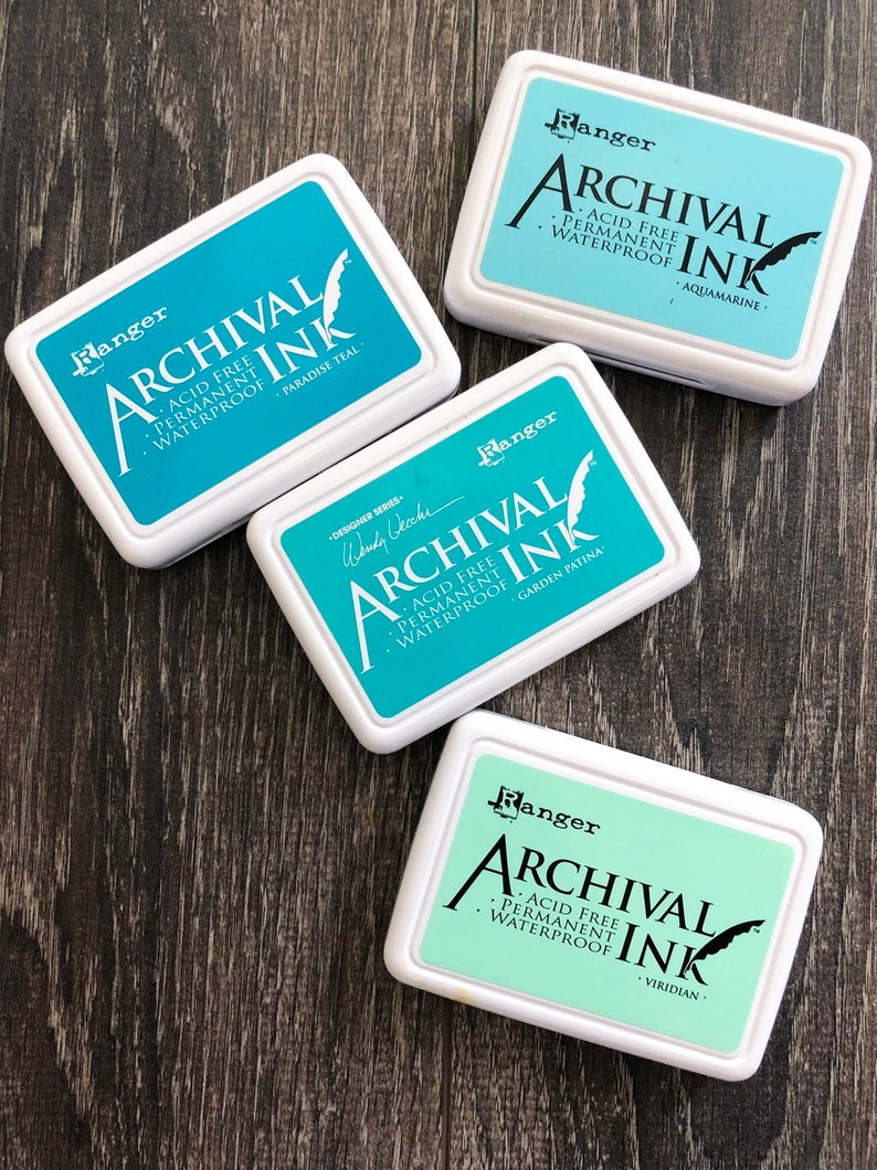 Peut inclure: Quatre tampons d'encre avec des couvercles blancs et de l'encre bleue et verte. Les tampons d'encre sont &eacute;tiquet&eacute;s "Ranger Archival Ink" et chacun a une couleur d'encre diff&eacute;rente : Paradise Teal, Aquamarine, Garden Patina et Viridian.