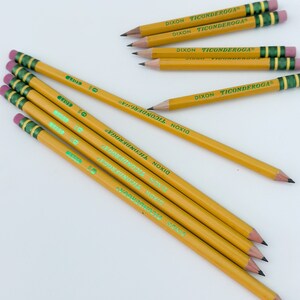 May include: A collection of yellow pencils with the text "DIXON TICONDEROGA" printed on the side. The pencils have green and gold bands near the eraser, and pink erasers. The pencils are arranged on a white surface.