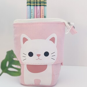 May include: A pink pencil case with a white cat design, holding several pencils. The pencils are pastel colors and have the text "Lindy King" printed on them. A single pink pencil rests in front of the case.