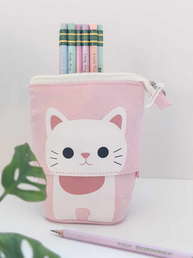 May include: A pink pencil case with a white cat design, holding several pencils. The pencils are pastel colors and have the text "Lindy King" printed on them. A single pink pencil rests in front of the case.