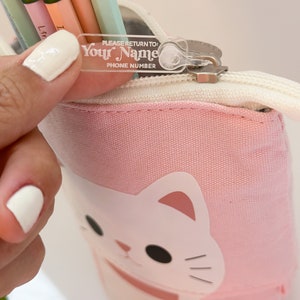 May include: Pink pencil case with a cartoon cat design. The zipper has a clear plastic tag that says "PLEASE RETURN TO: YOUR NAME PHONE NUMBER".
