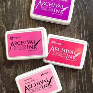 Peut inclure: Quatre tampons d'encre dans des contenants blancs avec des lettres noires. Les tampons d'encre sont &eacute;tiquet&eacute;s "Ranger Archival Ink" et chacun a une encre de couleur diff&eacute;rente : fleur de cactus, teinte magenta, fuchsia vibrant et pivoine rose.
