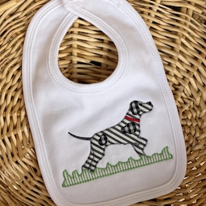 May include: White baby bib with a black and white patterned dog applique, a red collar, and green grass detail. The bib has a rounded shape with a neck opening and is set against a woven basket background.