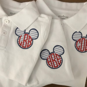 May include: Three white polo shirts with a Mickey Mouse-inspired design. The design features a blue and white striped head with a red monogram. Each shirt has a button-down collar and short sleeves.