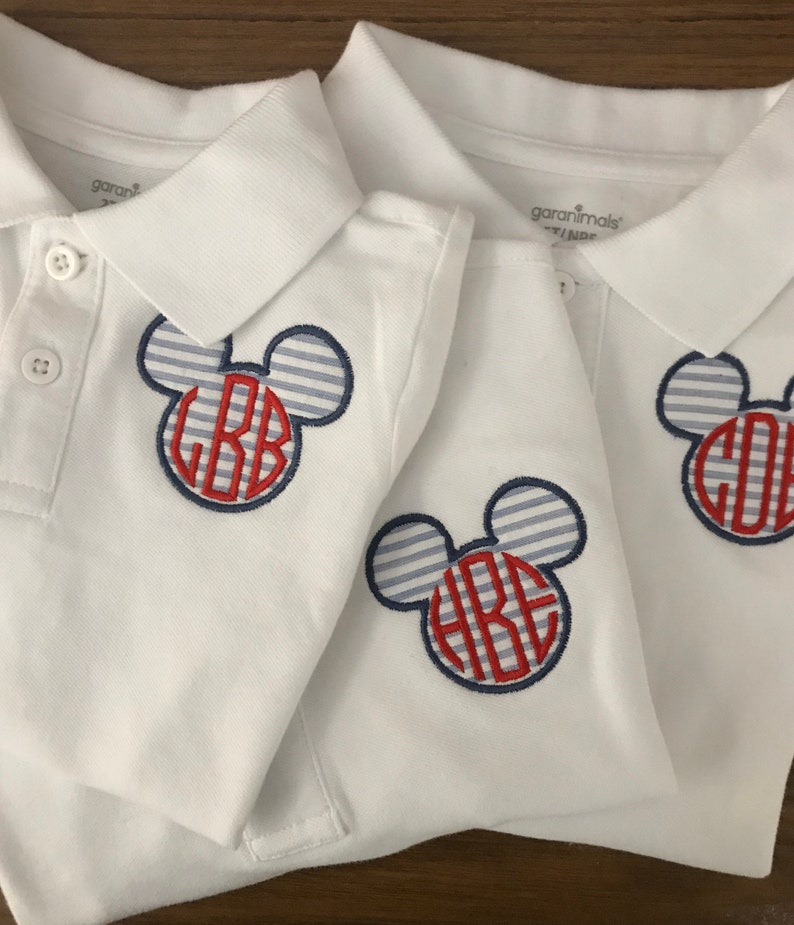 May include: Three white polo shirts with a Mickey Mouse-inspired design. The design features a blue and white striped head with a red monogram. Each shirt has a button-down collar and short sleeves.