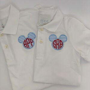 May include: Two white polo shirts with blue and red Mickey Mouse-shaped monograms. The monograms feature initials and are set against a blue and white striped background. The shirts have button-down collars and short sleeves.