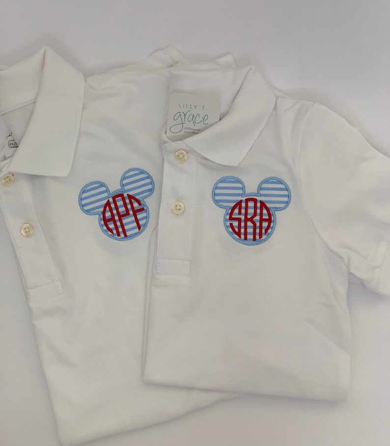 May include: Two white polo shirts with blue and red Mickey Mouse-shaped monograms. The monograms feature initials and are set against a blue and white striped background. The shirts have button-down collars and short sleeves.