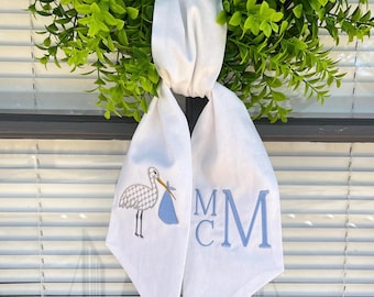 May include: White fabric door hanger with a blue and white stork carrying a baby. The fabric is embroidered with the monogram "MCM".