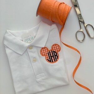 May include: White polo shirt with a Mickey Mouse-shaped patch in orange gingham with black initials. An orange ribbon spool and silver scissors are next to the shirt. The shirt has a tag that says "Lilly T. Grace".