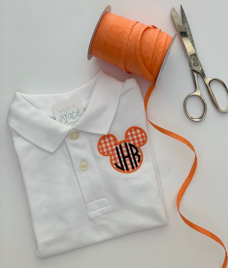 May include: White polo shirt with a Mickey Mouse-shaped patch in orange gingham with black initials. An orange ribbon spool and silver scissors are next to the shirt. The shirt has a tag that says "Lilly T. Grace".