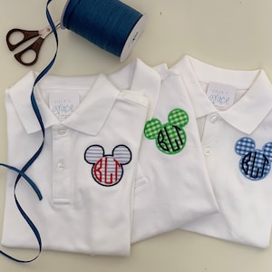 May include: Three white polo shirts with Mickey Mouse-shaped monograms. One has blue and red striped ears, another has green gingham ears, and the third has blue gingham ears. The shirts have the text "Lilly T. Grace" on the tag.
