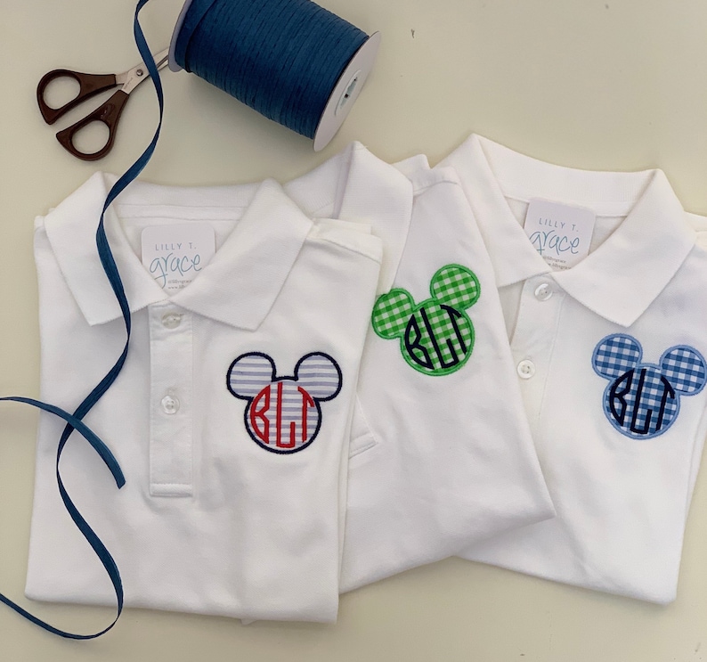 May include: Three white polo shirts with Mickey Mouse-shaped monograms. One has blue and red striped ears, another has green gingham ears, and the third has blue gingham ears. The shirts have the text "Lilly T. Grace" on the tag.