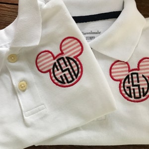 May include: Two white polo shirts with a Mickey Mouse-shaped design. The design features a red and white striped pattern with a black monogram. The shirts have a button-down collar and are labeled as size 3T/NP3.