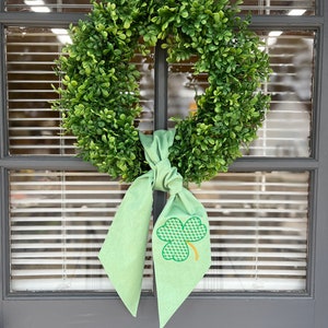 May include: A green wreath made of artificial boxwood with a green shamrock bow. The shamrock is embroidered with a gold outline.