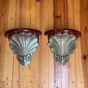 May include: Pair of antique wall shelves with a shell design. Each shelf has a dark red, semi-circular top and a gold-colored, fan-shaped support. The supports feature intricate scrollwork and a detailed, textured surface. The shelves are mounted on a wooden wall.