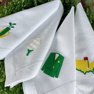 May include: White linen napkins with embroidered golf-themed designs. Each napkin features a different design: a golf bag, a golf ball on a tee, a green blazer, and a map of a golf course with a flag.