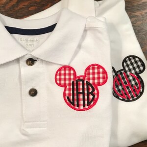 May include: Two white polo shirts with a button-down collar. One shirt features a red and white gingham Mickey Mouse head with the initials "JAB" in black. The other shirt has a black and white gingham Mickey Mouse head with the initials "JAH".