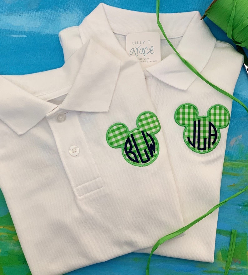 May include: Two white polo shirts with green and white gingham Mickey Mouse head patches. Each patch has a monogram in navy blue. The shirts have a button-down collar and the tag reads "Lilly T. Grace".