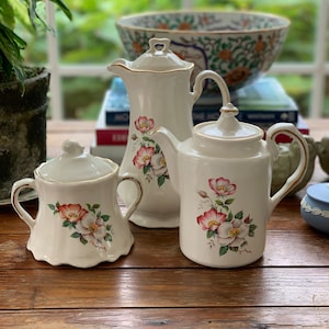 May include: A white ceramic tea set with a floral design. The set includes a teapot, creamer, and sugar bowl. The flowers are pink and white with green leaves.