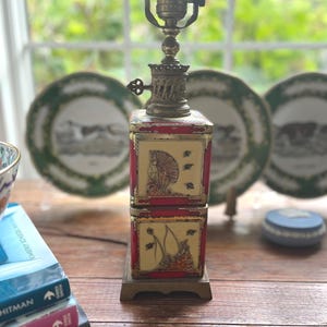 May include: A table lamp featuring a square, stacked tin base with red and cream panels. The panels display illustrations of figures and sailboats. The lamp has a brass-colored metal socket and a decorative, ornate metal base and top section.