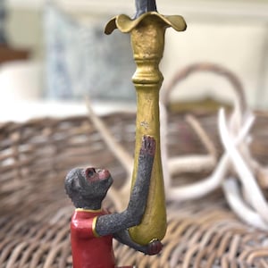 May include: A decorative candle holder featuring a monkey figurine in a red outfit with gold trim, holding a gold-colored candle holder. The candle holder has a dark gray, textured candle. The figurine is set on a woven wicker surface.