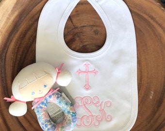 May include: White bib with pink embroidered cross and monogram. A pink and blue floral baby rattle is in the foreground.