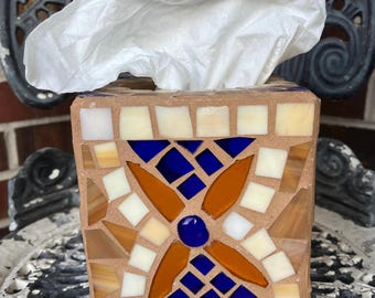 May include: A square tissue box with a mosaic tile design. The mosaic features a central flower pattern in shades of blue, orange, and white. The box is filled with white tissues, with some extending from the top.