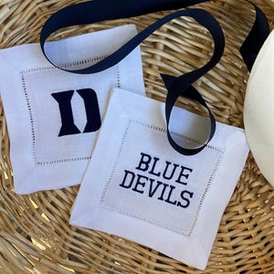 May include: Two white linen coasters with navy blue embroidered text. One coaster features the letter "D", and the other reads "BLUE DEVILS". A navy blue ribbon is draped across the coasters, which are set in a woven basket.