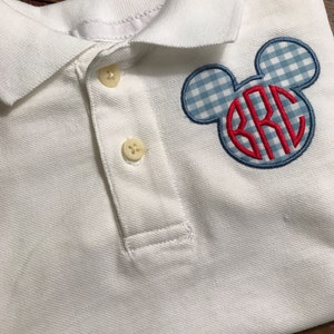 May include: White polo shirt with a blue and white gingham Mickey Mouse head design. The center of the design features red embroidered initials. The shirt has a classic collar and two buttons.