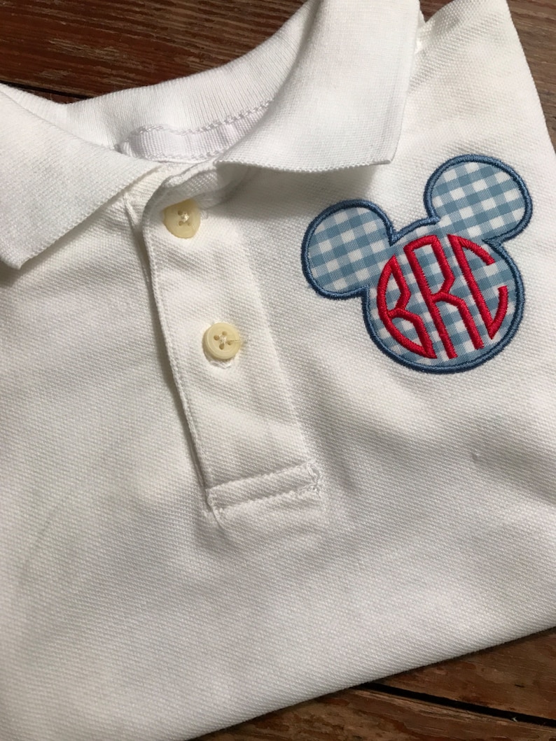 May include: White polo shirt with a blue and white gingham Mickey Mouse head design. The center of the design features red embroidered initials. The shirt has a classic collar and two buttons.