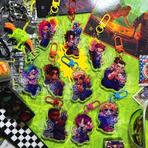 May include: A selection of acrylic keyrings featuring anime-style characters in various poses. The keyrings have colourful designs and are attached to metal clasps in various colours. The background is a bright green surface with other objects.