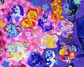 Könnte beinhalten: Eine Sammlung farbenfroher Aufkleber zum Thema Ponys mit verschiedenen Designs, darunter Einhörner und Pegasus. Die Aufkleber weisen leuchtende Farben wie Blau, Rosa, Gelb und Lila auf. Einige Aufkleber haben Glitzerakzente. Die Aufkleber sind auf einer rosa-lila karierten Oberfläche angeordnet.