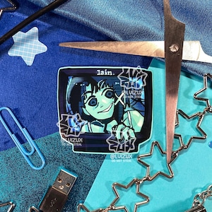 May include: A blue and black sticker featuring a cartoon character with large eyes and a creepy smile. The character is looking out of a television screen with the word "lain." printed above. The sticker has a white border and a copyright notice at the bottom.