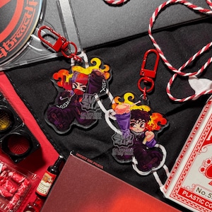 Può includere: Due portachiavi in acrilico con chiusure a moschettone rosse. Ogni portachiavi presenta un personaggio dei cartoni animati con fiamme arancioni e gialle, che indossa abiti viola e neri. Il testo "CLVIZUX DO NOT STEAL" è visibile.