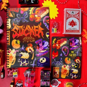 Könnte beinhalten: Ein Spiralnotizbuch und Stickerbögen mit farbenfrohen Illustrationen und dem Wort "STICKER" in fetter Schrift. Ein Kartenspiel, ein gelber Stern und andere kleine Gegenstände sind auf einer roten Oberfläche angeordnet.