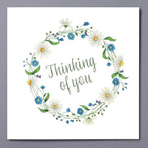 May include: A white card featuring a floral wreath. The wreath is made up of white daisies with yellow centres and small blue flowers, set on green stems. The words "Thinking of you" are written in green script.