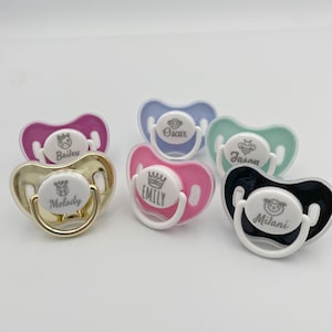 May include: Six personalized pacifiers with different colors and designs. The pacifiers are white with a variety of colors and designs. The pacifiers are personalized with the names "Baileu", "Melody", "Emily", "Oscar", "Jason", and "Milani".
