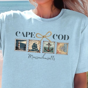 Op de afbeelding: Lichtblauw T-shirt met een Cape Cod-ontwerp. Het ontwerp bevat de woorden "Cape Cod Massachusetts" en vier postzegelachtige afbeeldingen van een vuurtoren, een schip, een anker en een kompas. Het shirt is gemaakt van zacht materiaal.