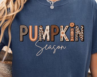 May include: A navy blue t-shirt with the words "PUMPKIN Season" in a mix of patterned fonts. The word "PUMPKIN" features a plaid, leopard print, and floral design. The word "Season" is written in a cursive font.