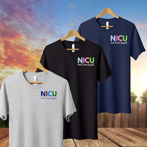 May include: Three t-shirts on wooden hangers. The shirts are gray, black, and navy blue. Each shirt has the word "NICU" in colorful letters, with "Saint Francis Hospital" below it in a script font. The background is a blurred sky and wooden deck.